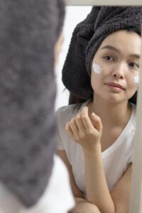 Woman applying skincare cream on her face while looking in a mirror after shower